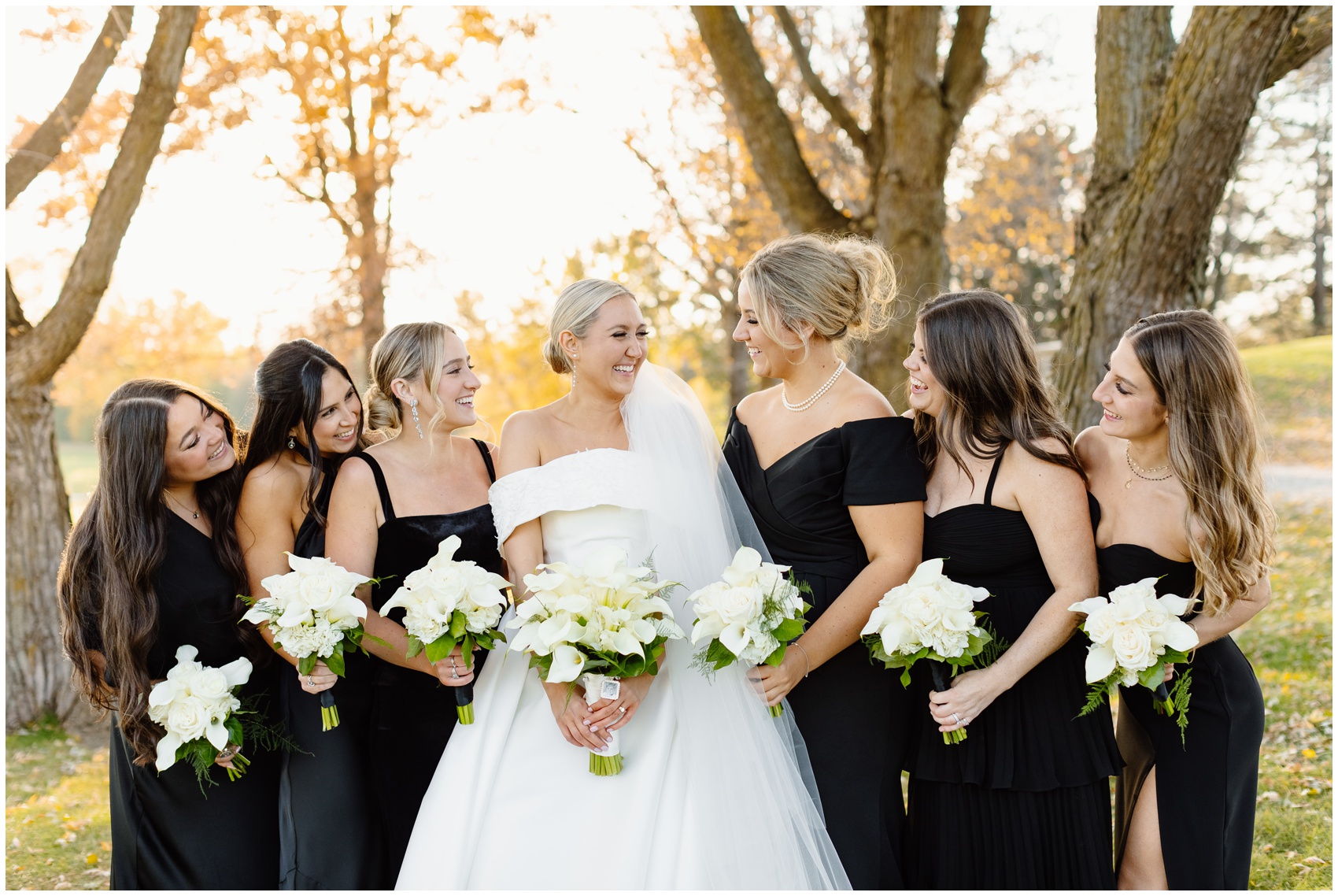 A bride laughs while standing with her bridesmaids in black dresses with white flowers at her Hyatt Lodge Oak Brook Wedding