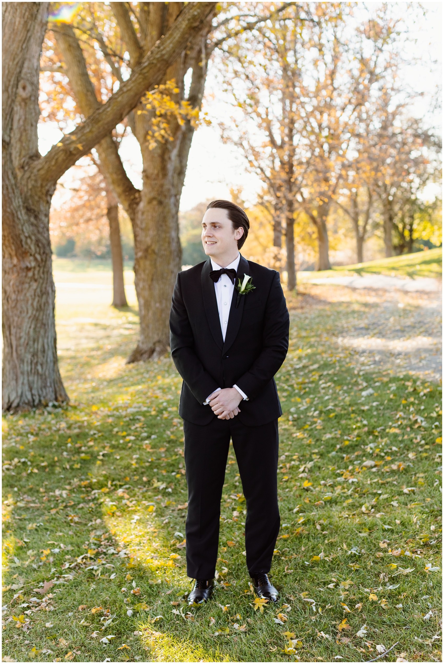 A groom stands with hands folded in front in a black tux under a tree at sunset