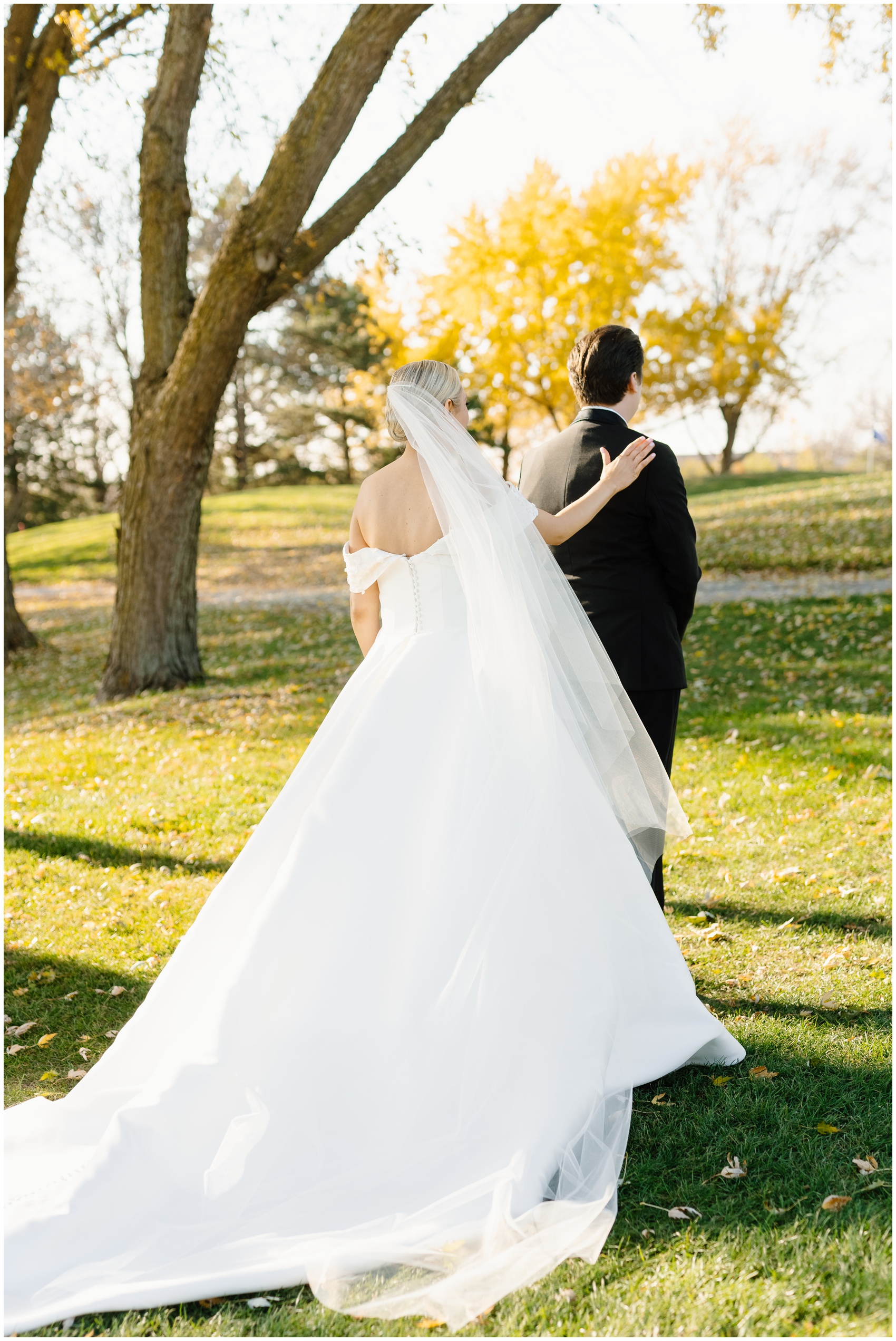 A bride with a long train taps the shoulder of her groom during their first look in a park lawn