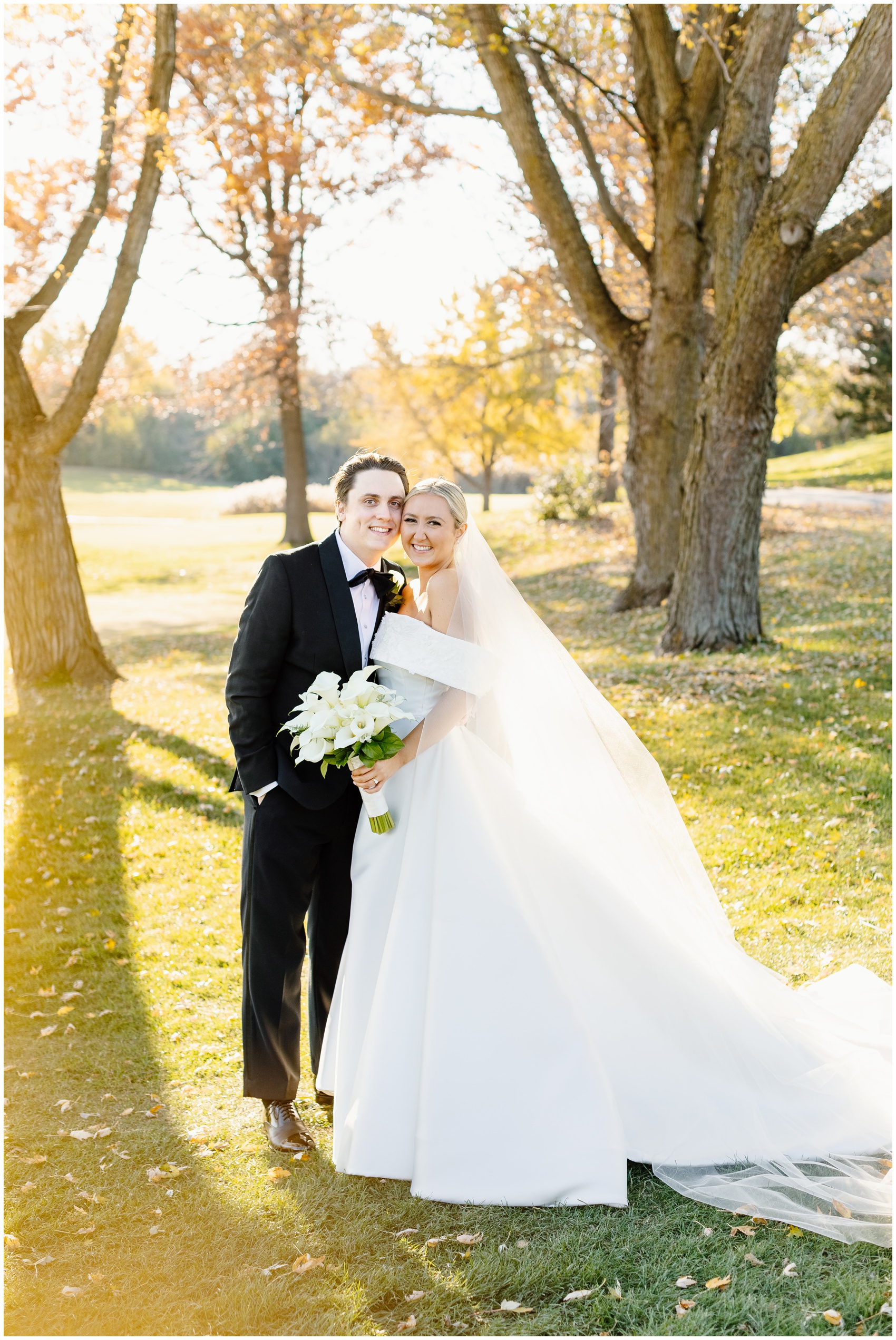 A bride and groom smile together while standing close in a park lawn at sunset