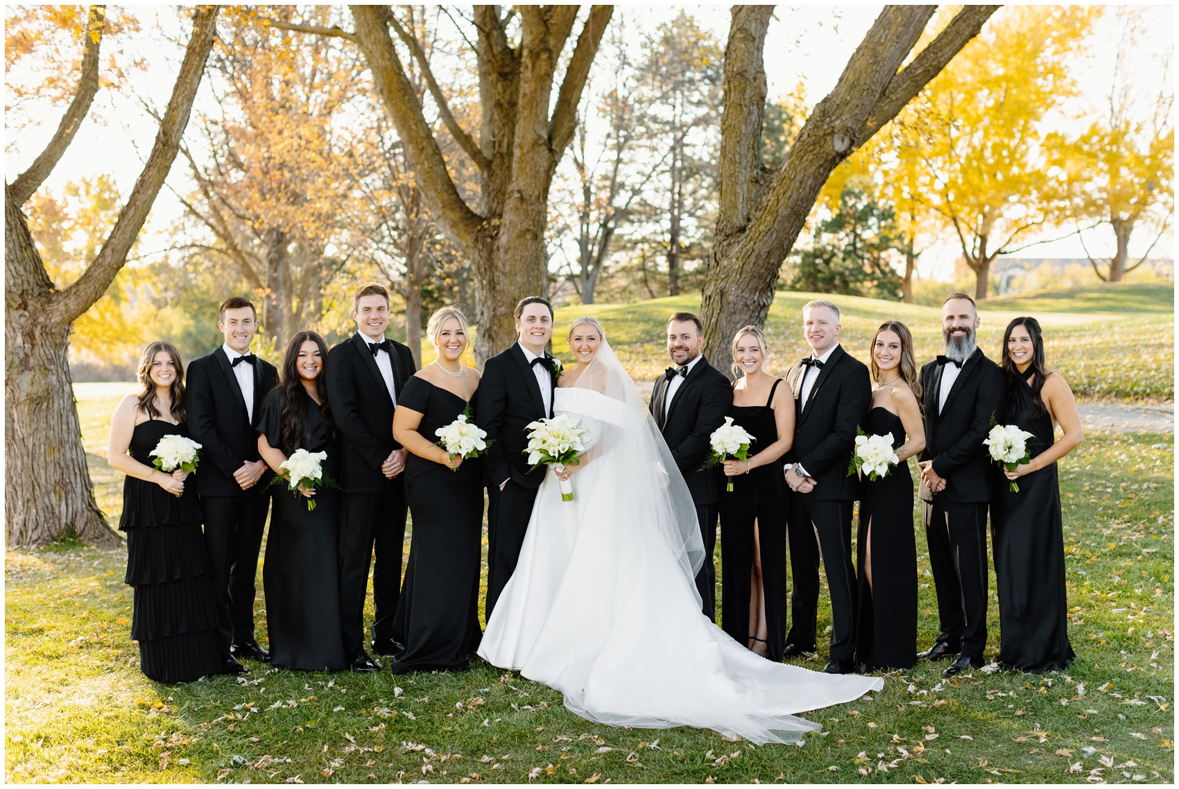 Newlyweds stand in a park lawn at sunset with their wedding party in all black