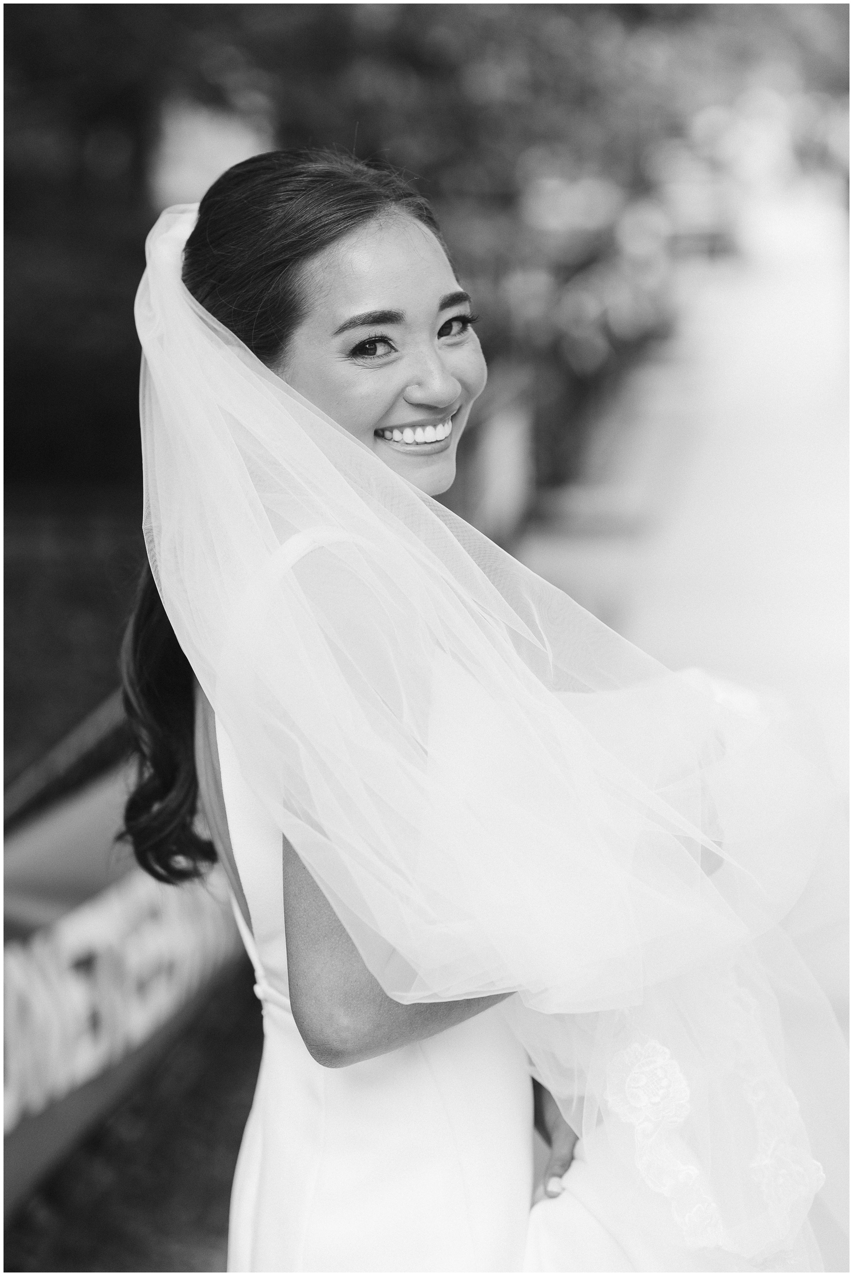 A bride smiles over her shoulder while walking on a sidewalk in black and white