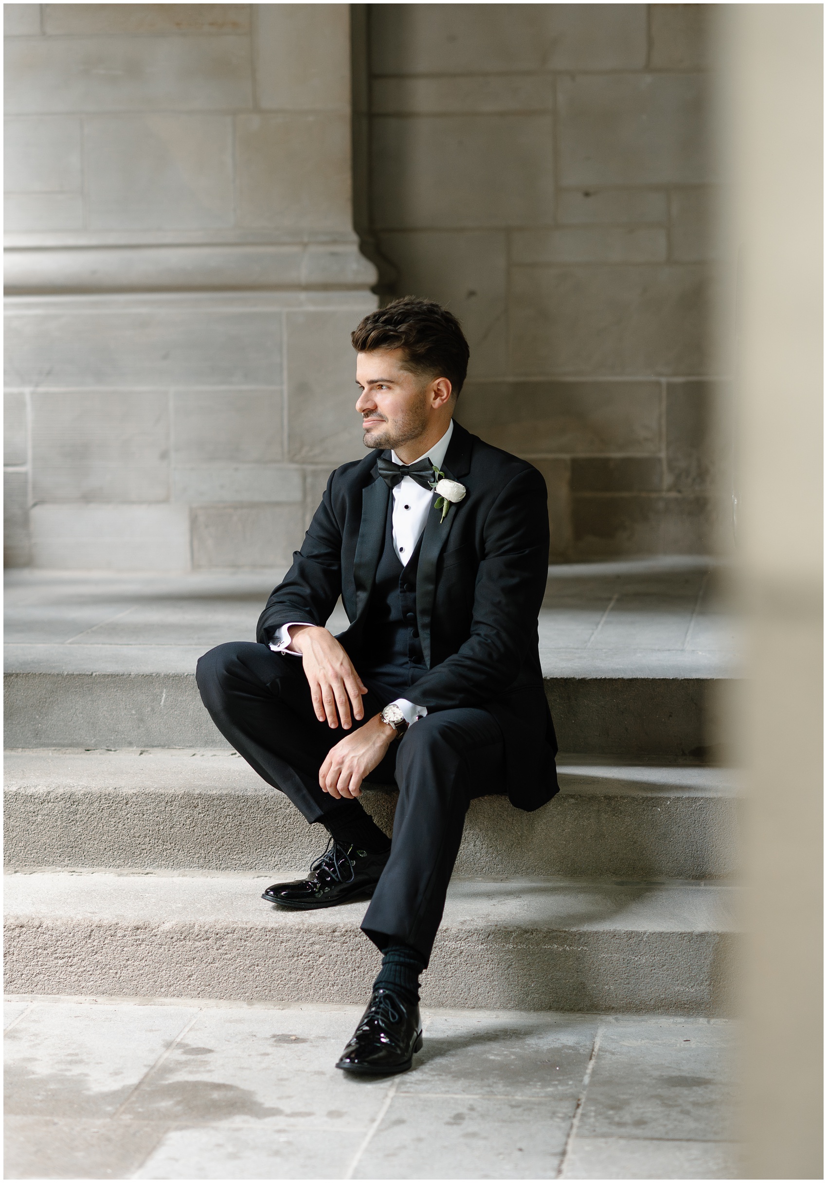 A groom sits on some steps smiling off into the distance in a black suit at the arbory chicago