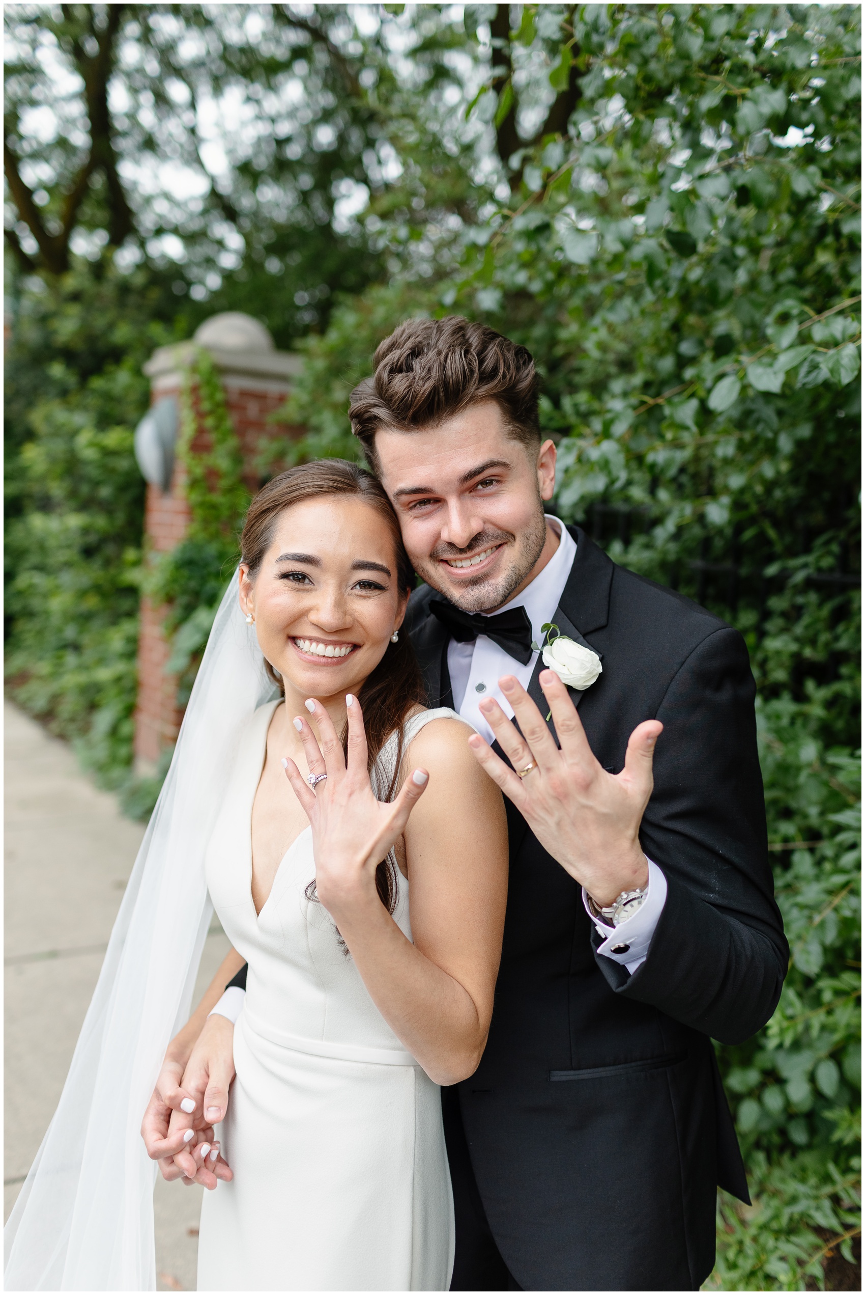 Newlyweds show off their rings while walking on a garden sidewalk