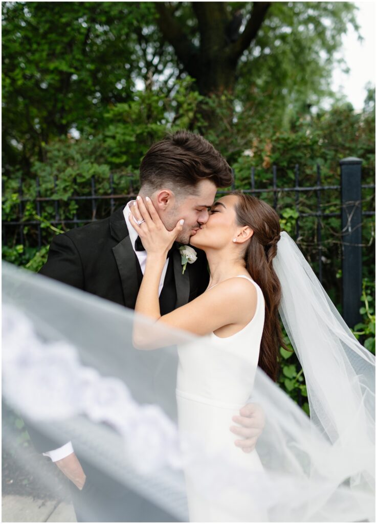 Newlyweds kiss as the veil flies around them by an iron fence