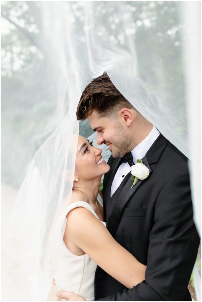 A bride and groom smile at each other while hugging under a large veil