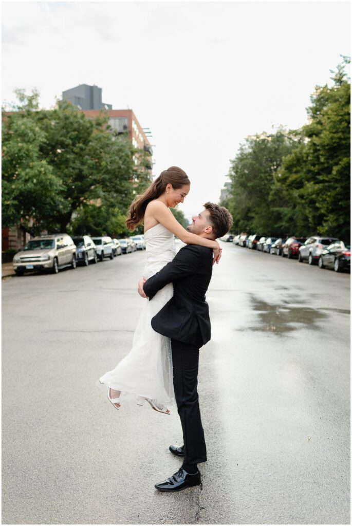 A groom in a black suit lifts his bride in the middle of a wet street during their wedding at the arbory chicago