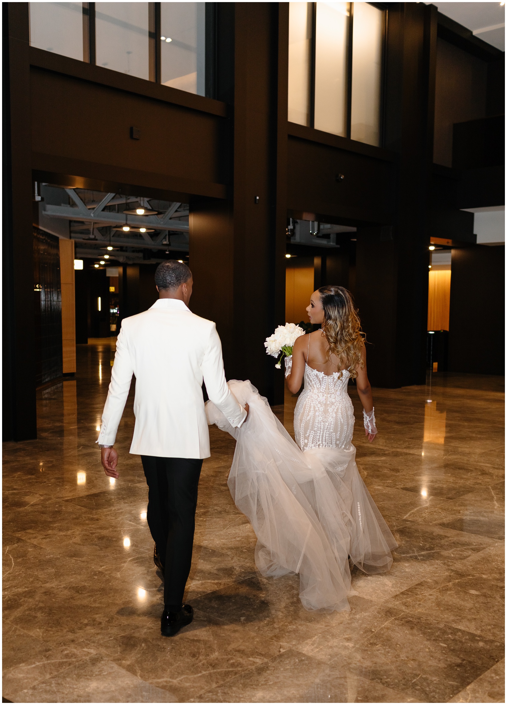 A groom holds the train of his bride as they walk together through a large lobby