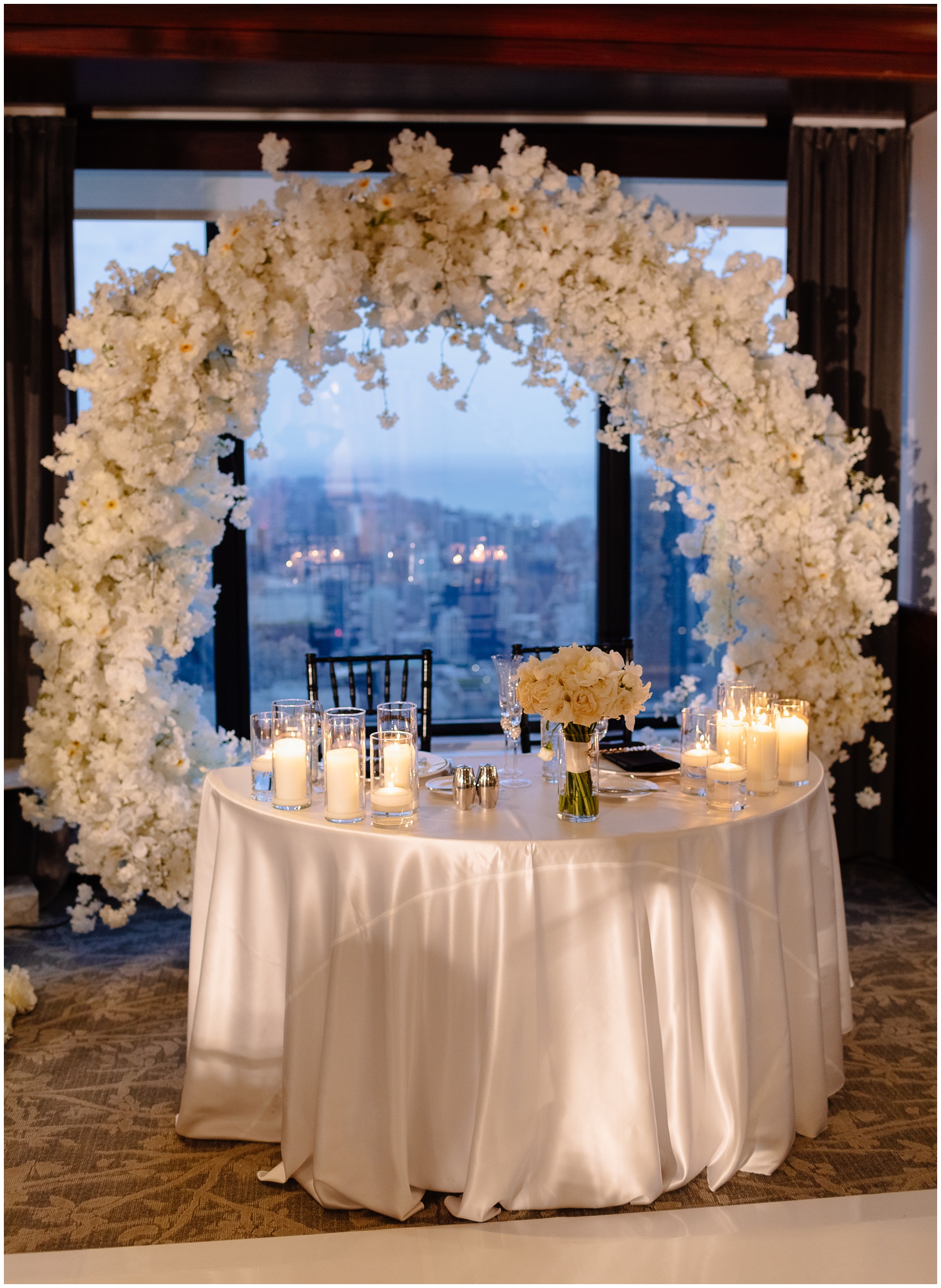 A view of a newlywed head table with a large white flower arch in a window designed by Chicago Wedding Planners