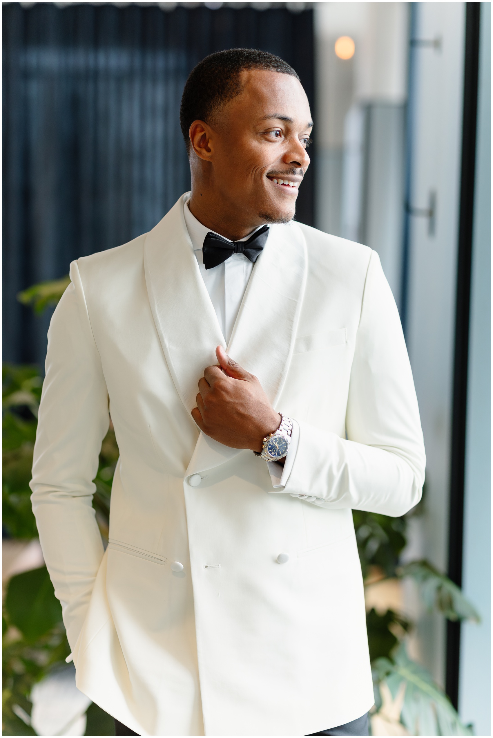 A groom smiles out a window in his white tuxedo while holding his lapel