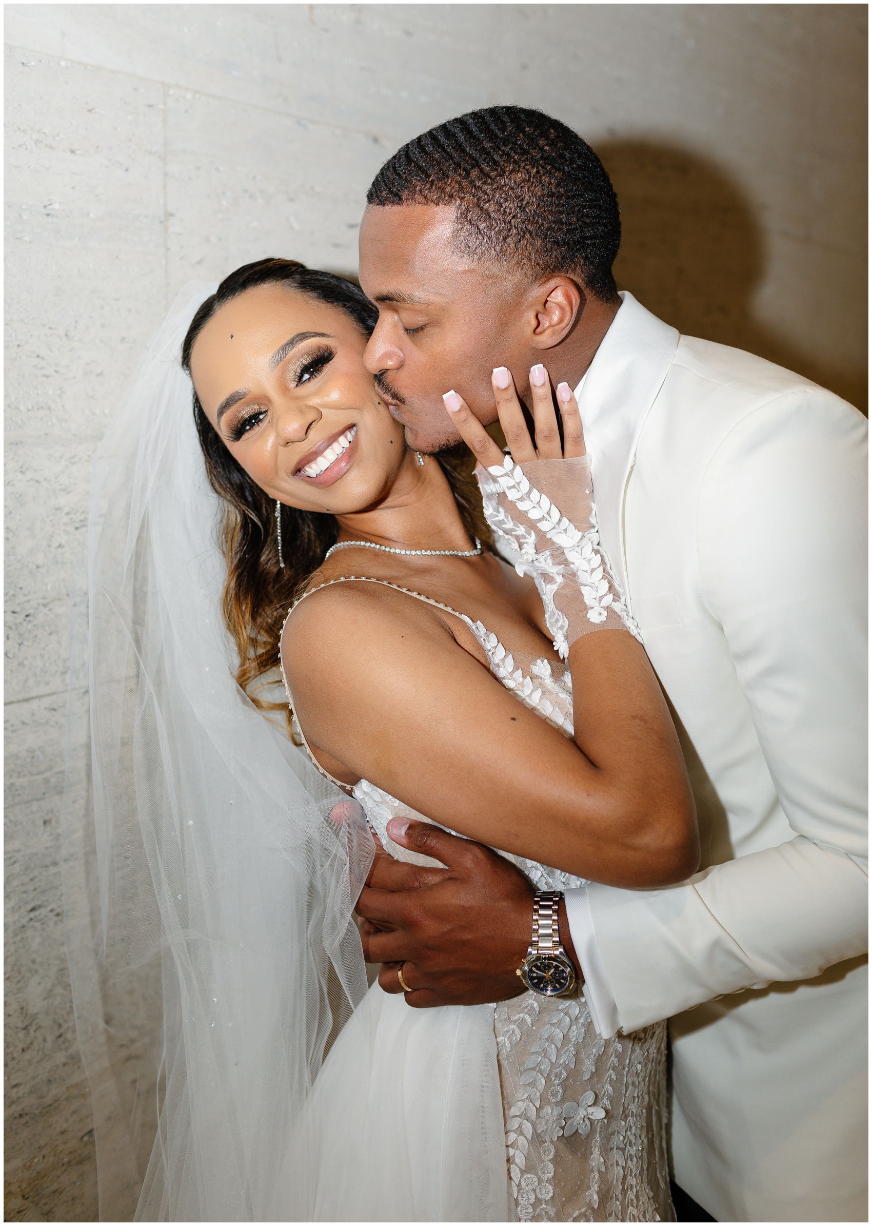 A bride smiles big while her husband kisses her cheek