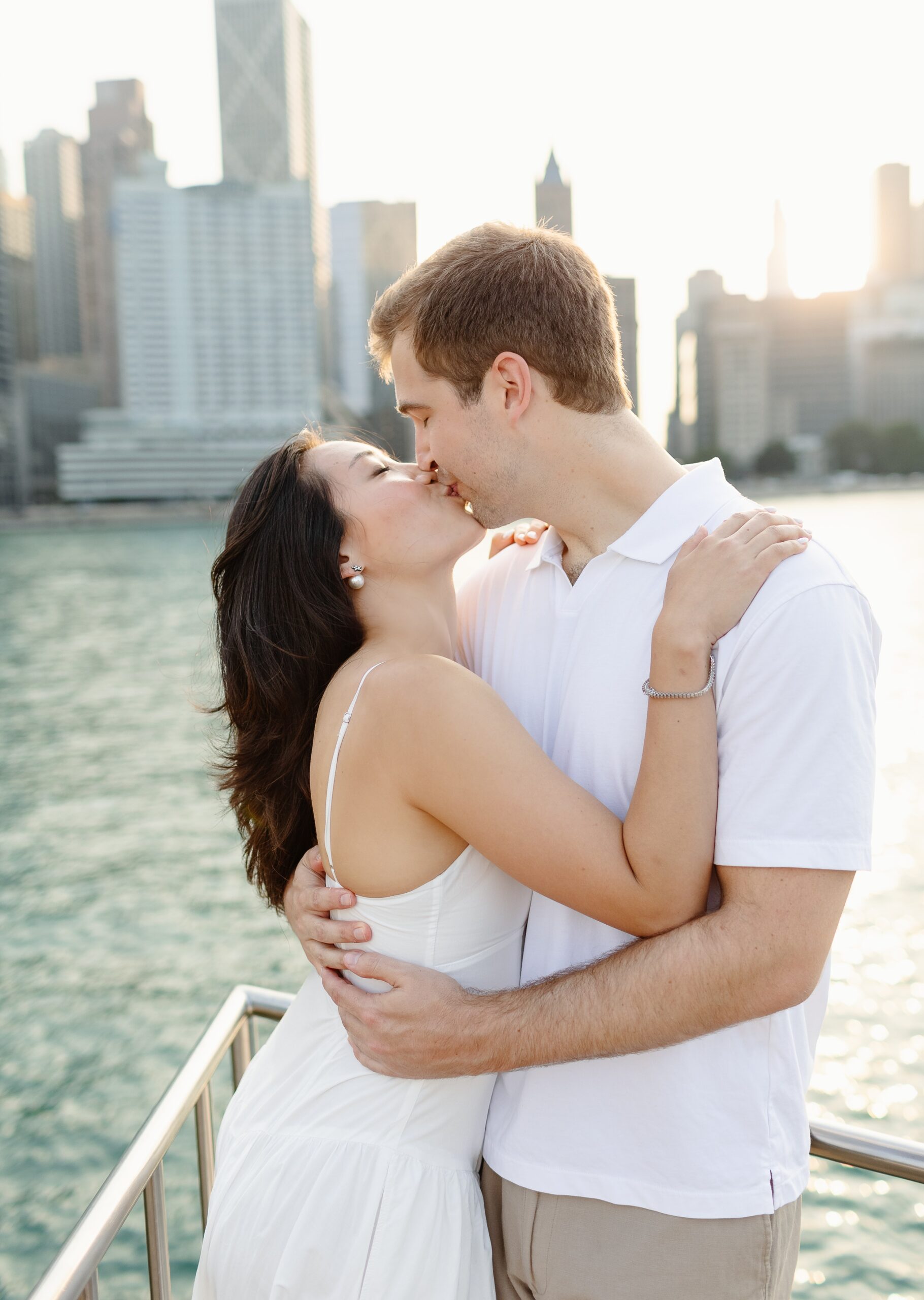 A couple kisses in white dress and shirt on a balcony overlooking the water at one of the engagement photo locations chicago