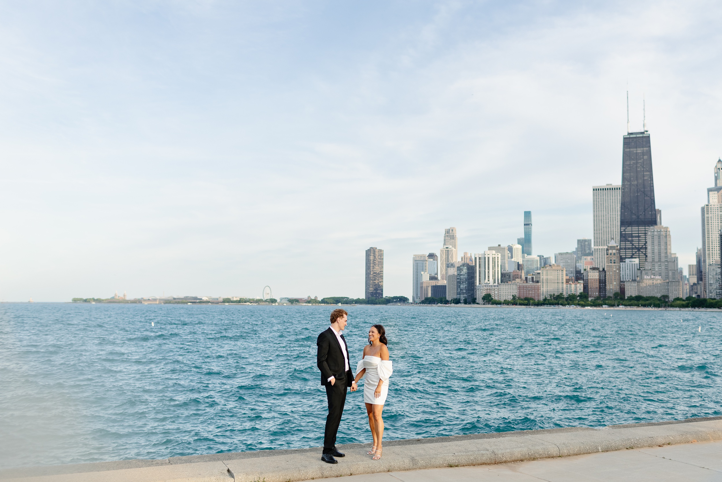 A happy engaged couple holds hands and laughs while walking on a lakeside park at sunset