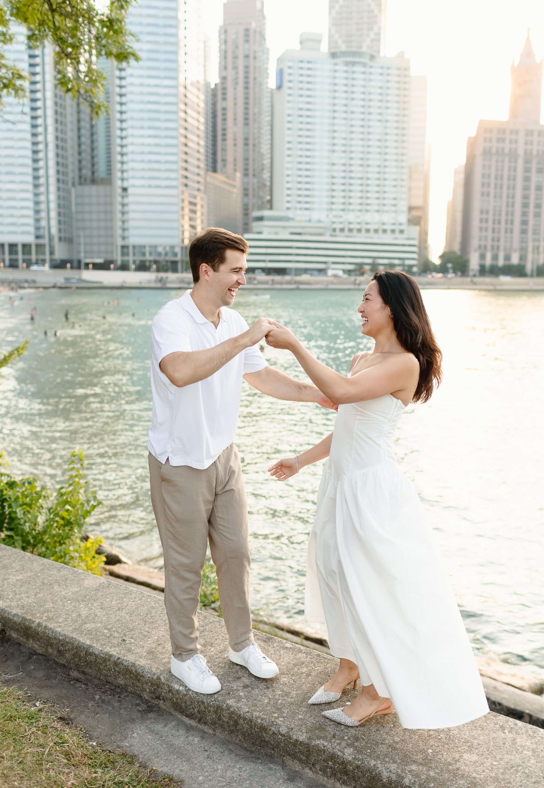 A happy couple laughs and dances on a wall above the water with the city behind them