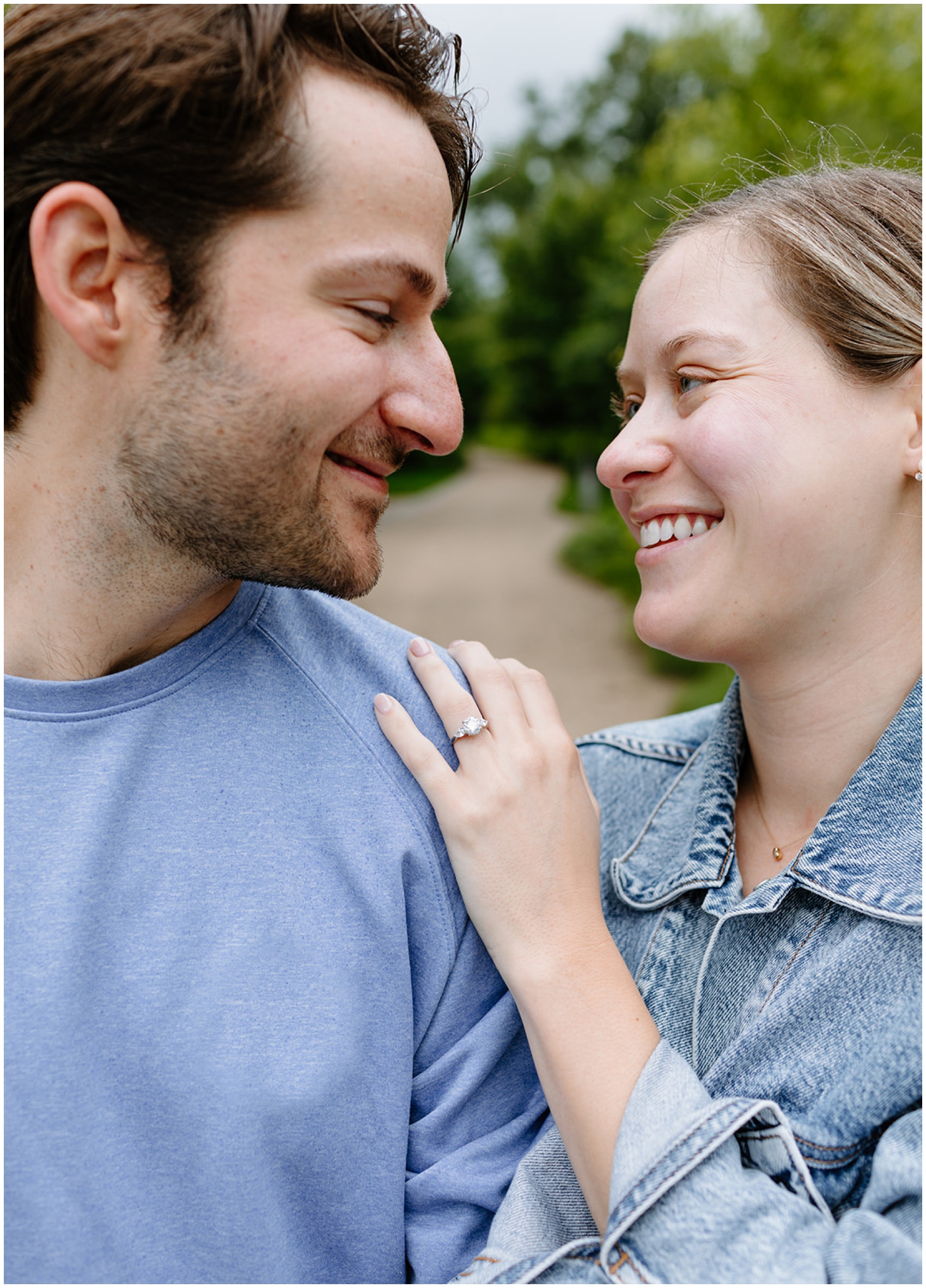 A woman in a denim jacket smiles and hangs on the arm of her fiancee as they explore a garden