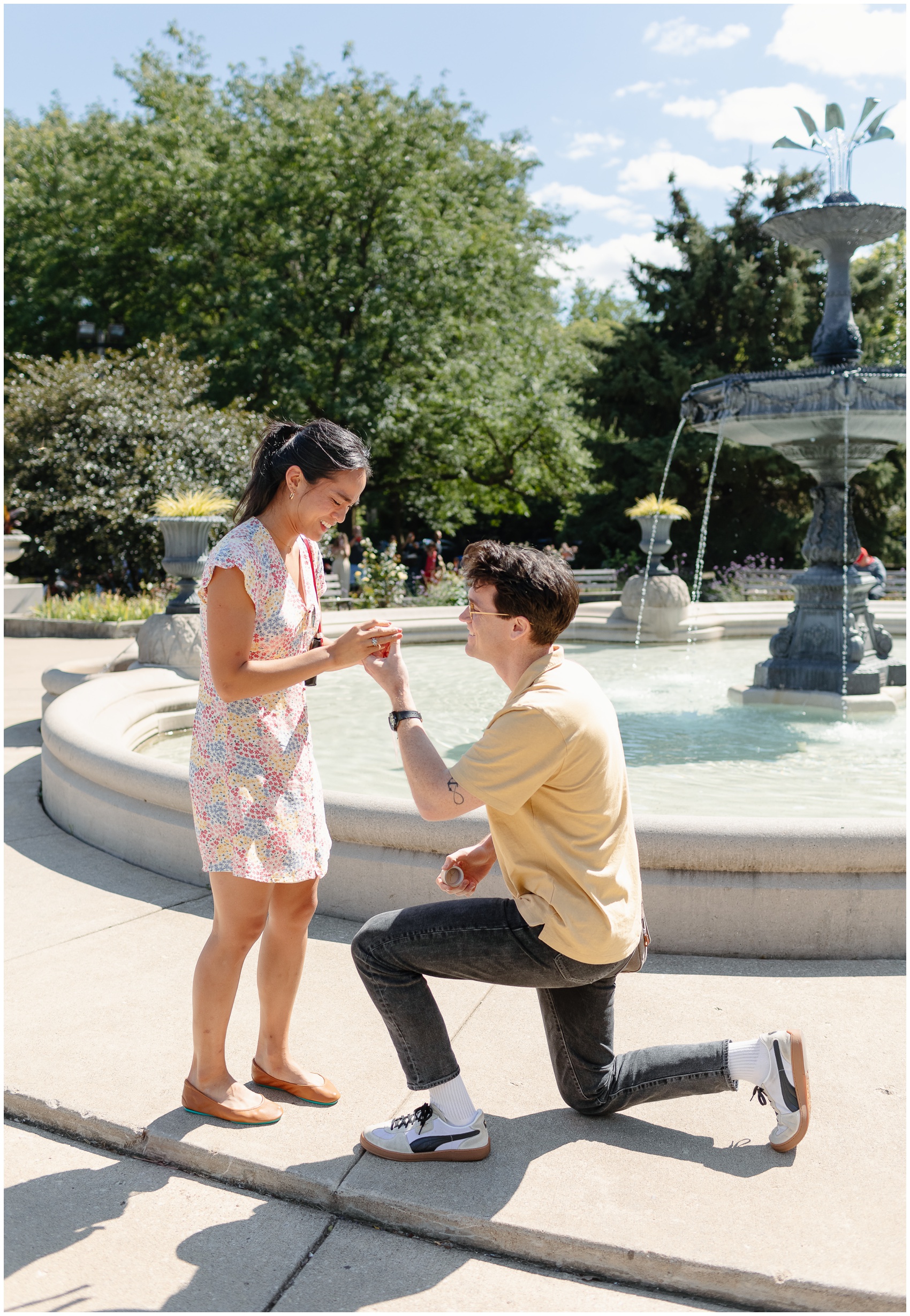 A man in a yellow shirt gets down on one knee while Proposing In Chicago in front of a large park fountain to his girlfriend in a colorful dress at mid day