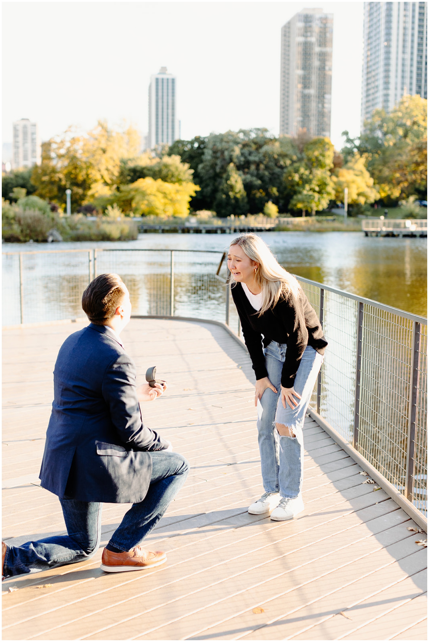 A man in a suit kneels on a dock while Proposing In Chicago to his ecstatic fiancee with hands on her knees