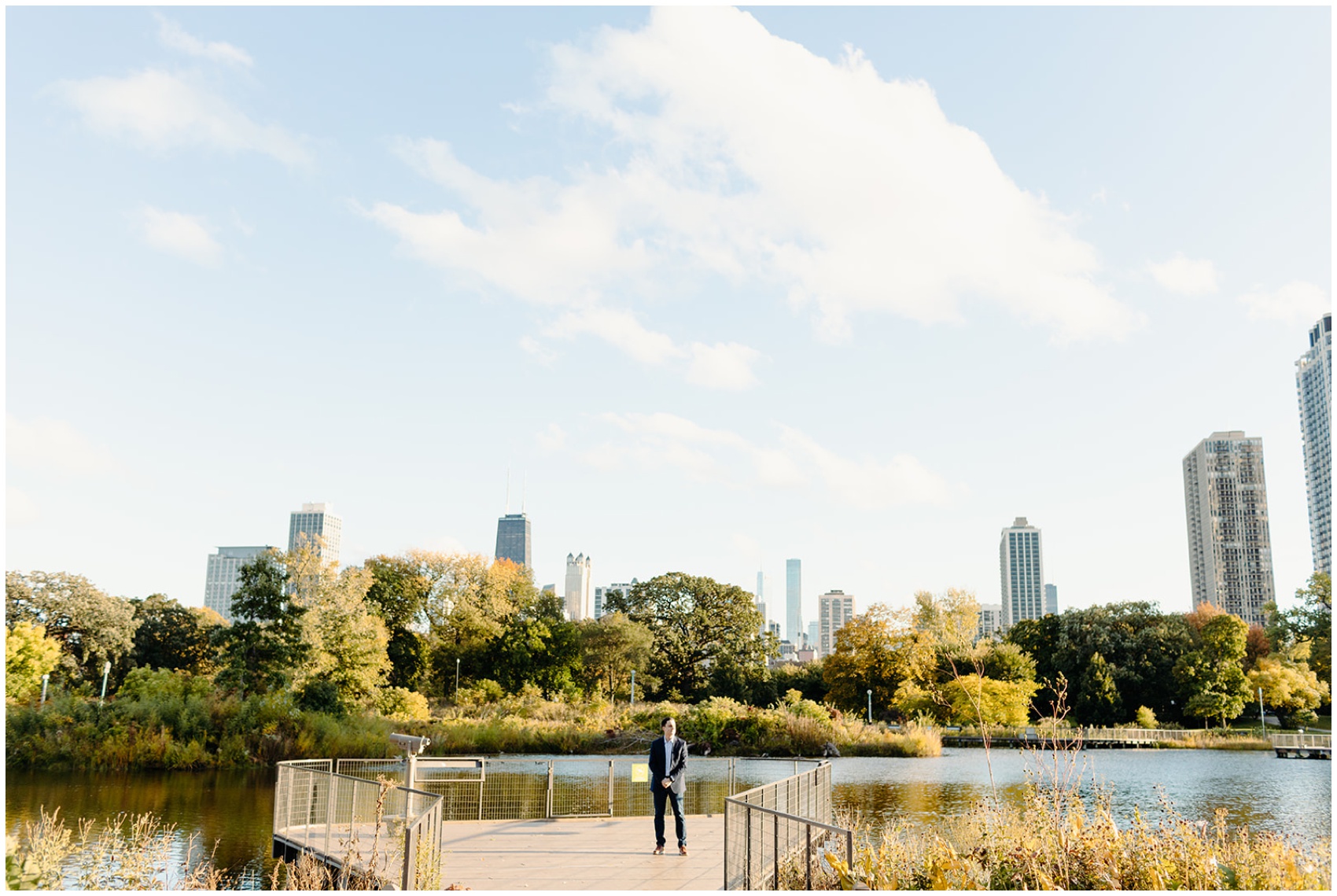 A man in a suit stands on a dock with the skyline behind him waiting for his girlfriend to propose