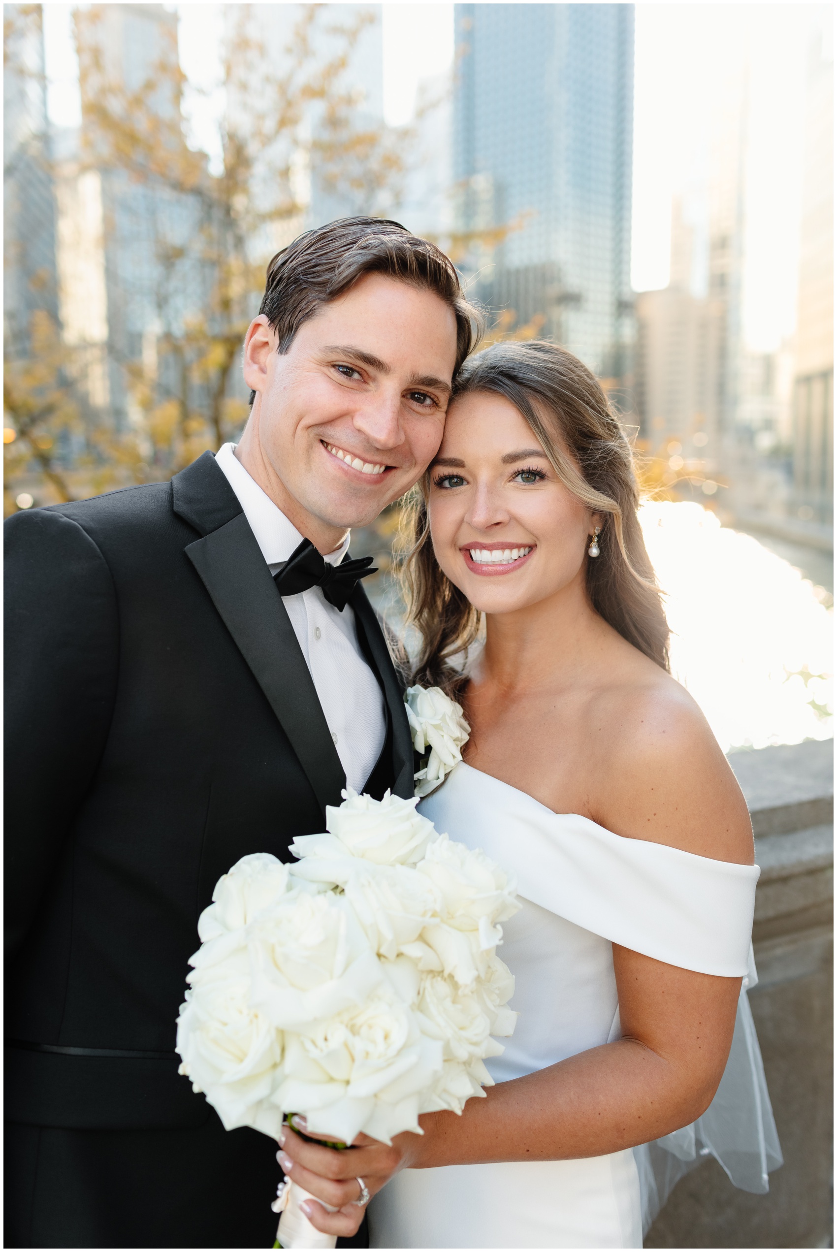 A bride and groom stand cheek to cheek on a bridge overlooking the river holding a large white bouquet