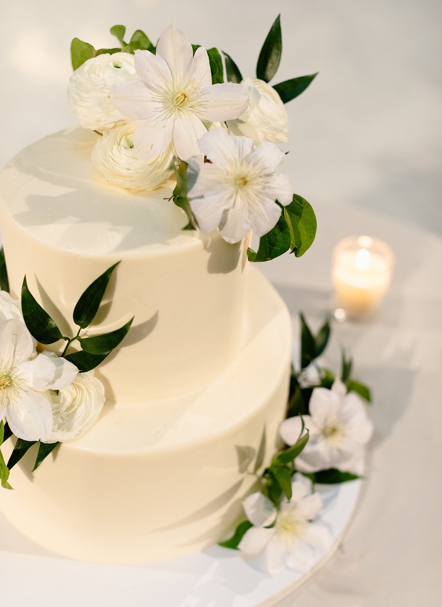 A two tier cake sits on a table with a candle covered in white flowers