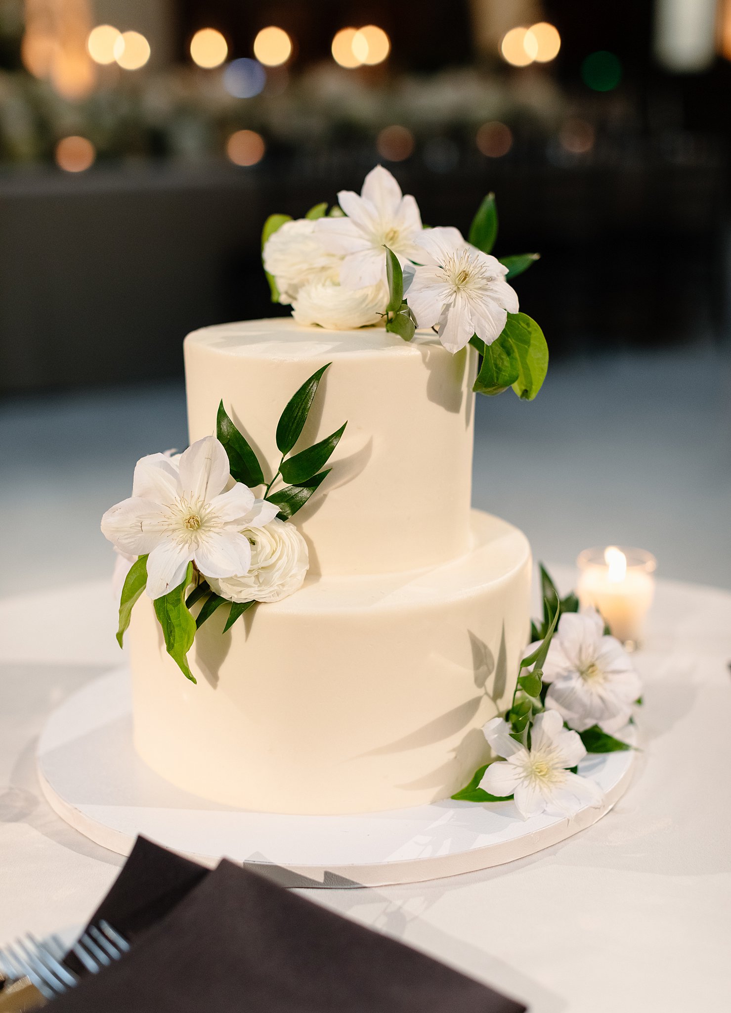 Details of a white two tier wedding cake with flowers sitting on a white linen table at a reception