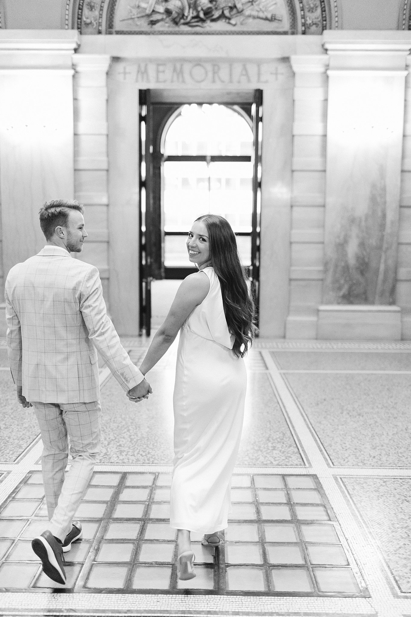 A woman smiles over her shoulder while walking out a historic lobby holding hands with her fiancee