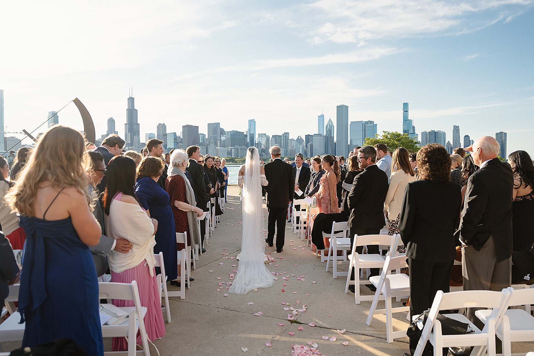 A bride walks down the aisle with dad over pink rose petals at an outdoor waterfront park wedding ceremony