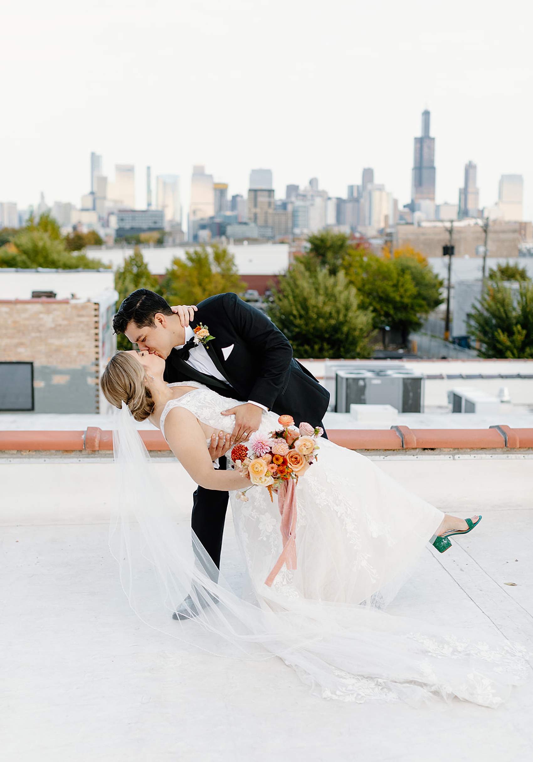 A groom in a black tuxedo dips his bride for a kiss on a rooftop