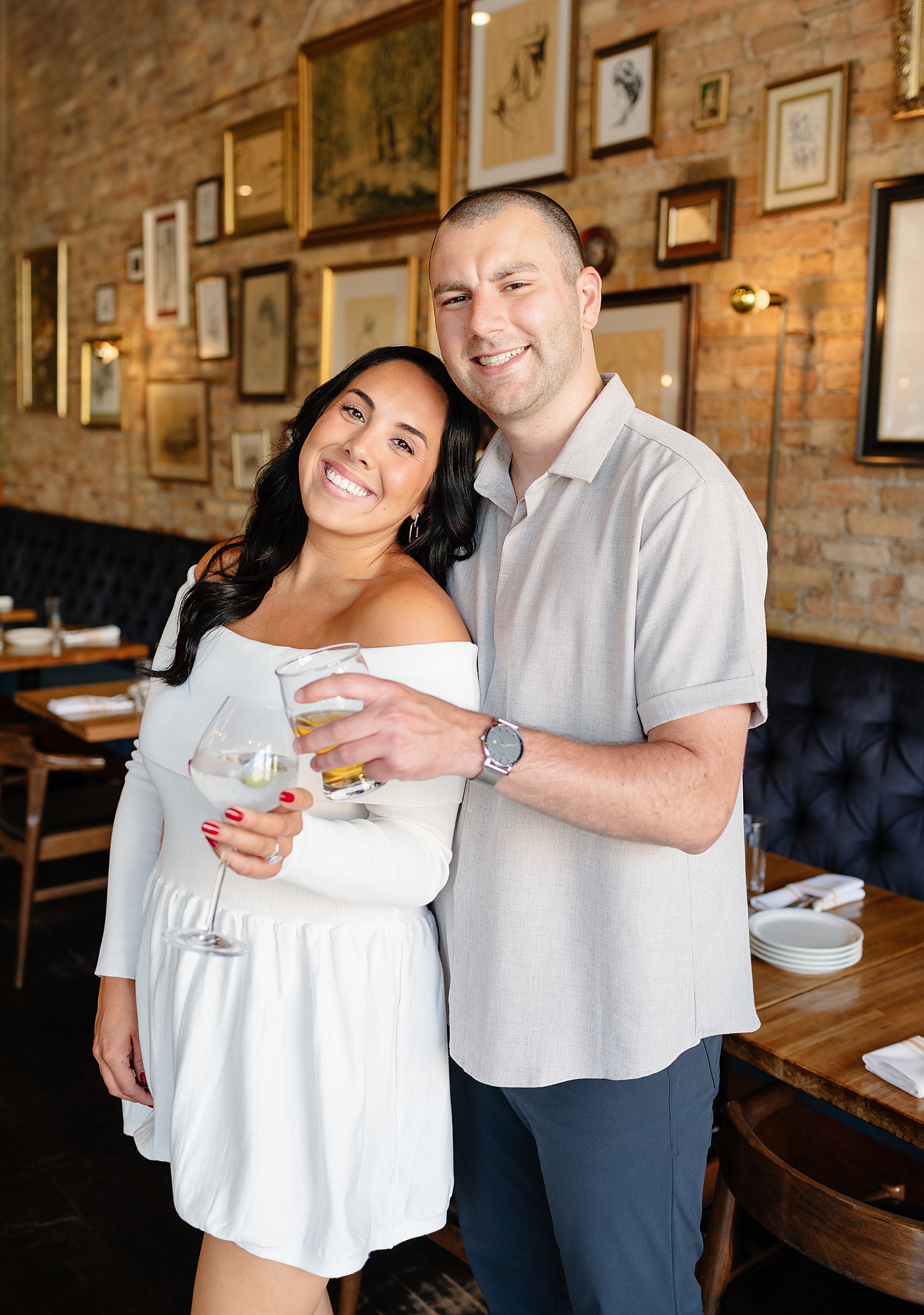A woman in white leans into her fiancee's chest as they toast drinks while standing in a bar