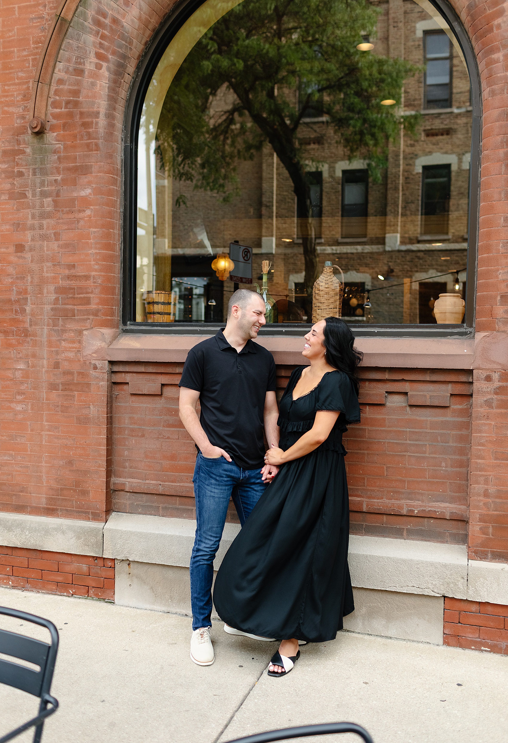 a happy couple hold hands and lean against a brick wall under a window on the sidewalk for their engagement photo ideas in chicago